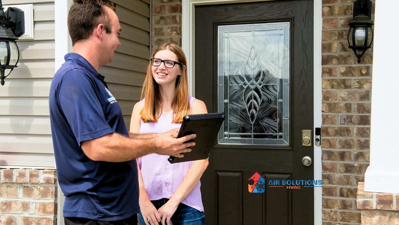 A professional in a blue polo holding a tablet speaks with a person outside a house with a dark brown front door.