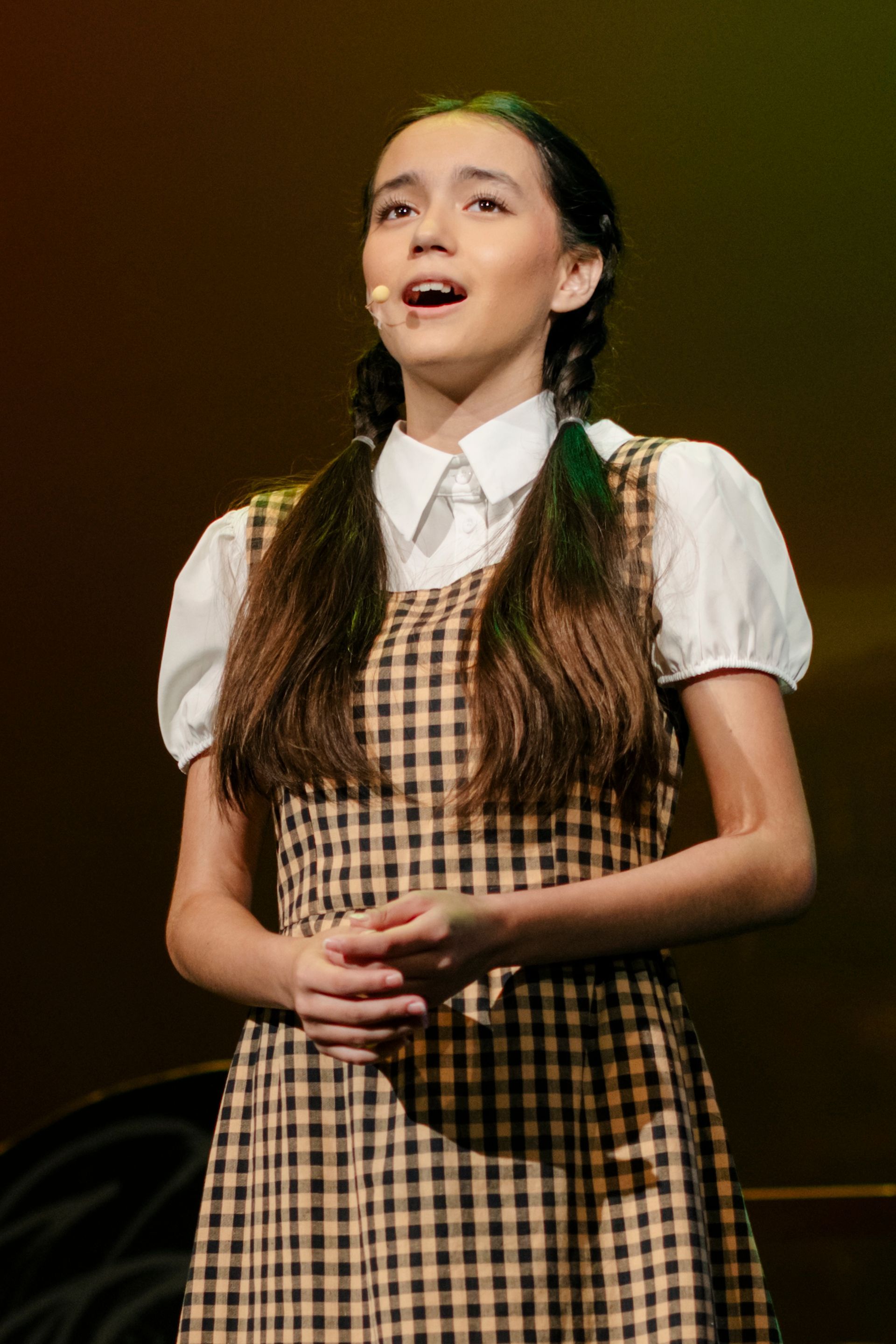Performer in a checkered dress and white blouse singing onstage under warm spotlight