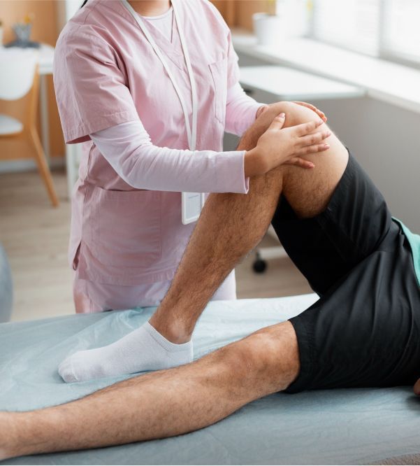 A person in pink scrubs examines a person's knee on a medical table, possibly physiotherapy.