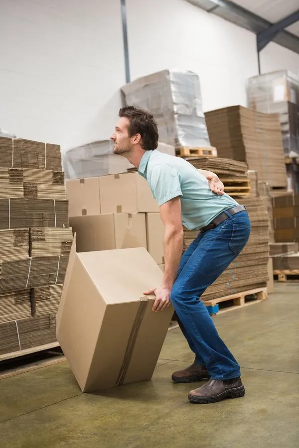 Man lifting a large cardboard box in a warehouse, back arched, showing a strained expression.