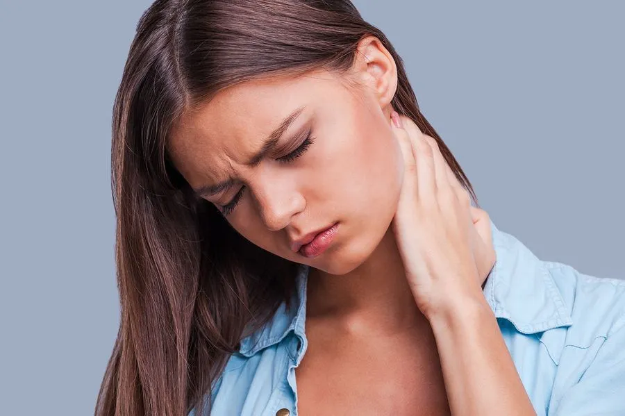 Woman with closed eyes, touching neck, likely in pain; blue shirt, gray background.