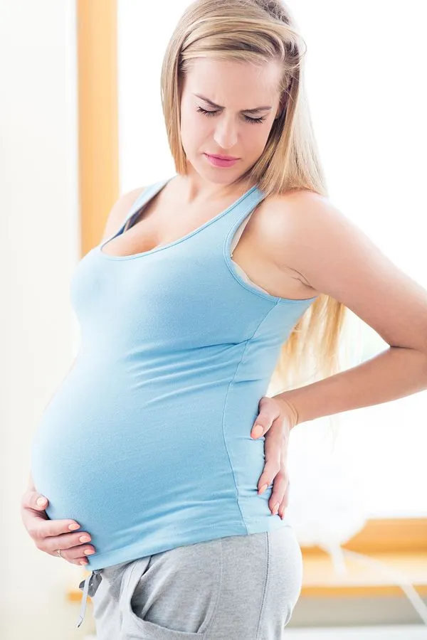 Pregnant woman, light blue top, gray pants, touching her lower back, appears to be in pain.