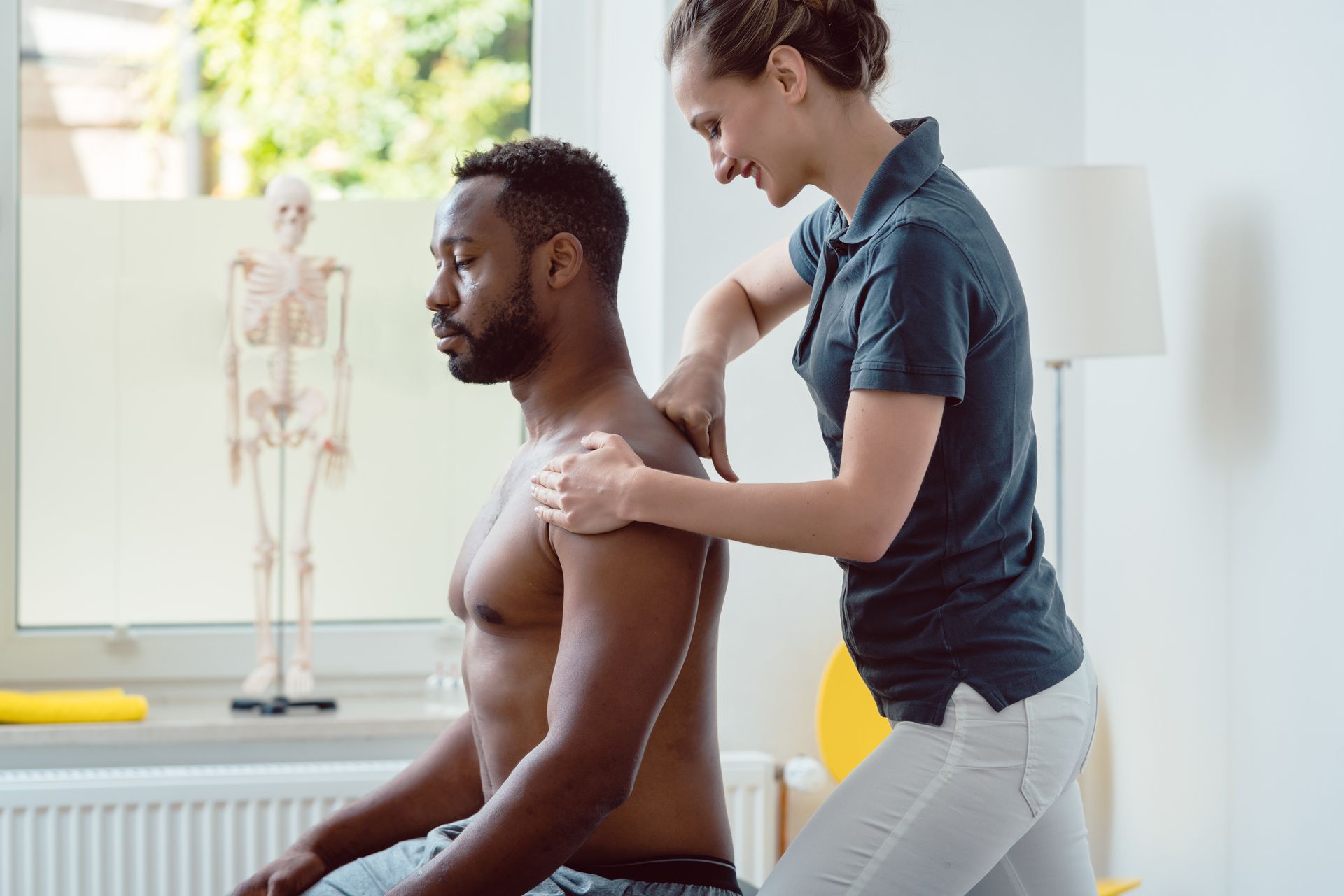 Woman giving a shoulder massage to a seated person; indoors with anatomical model in background.