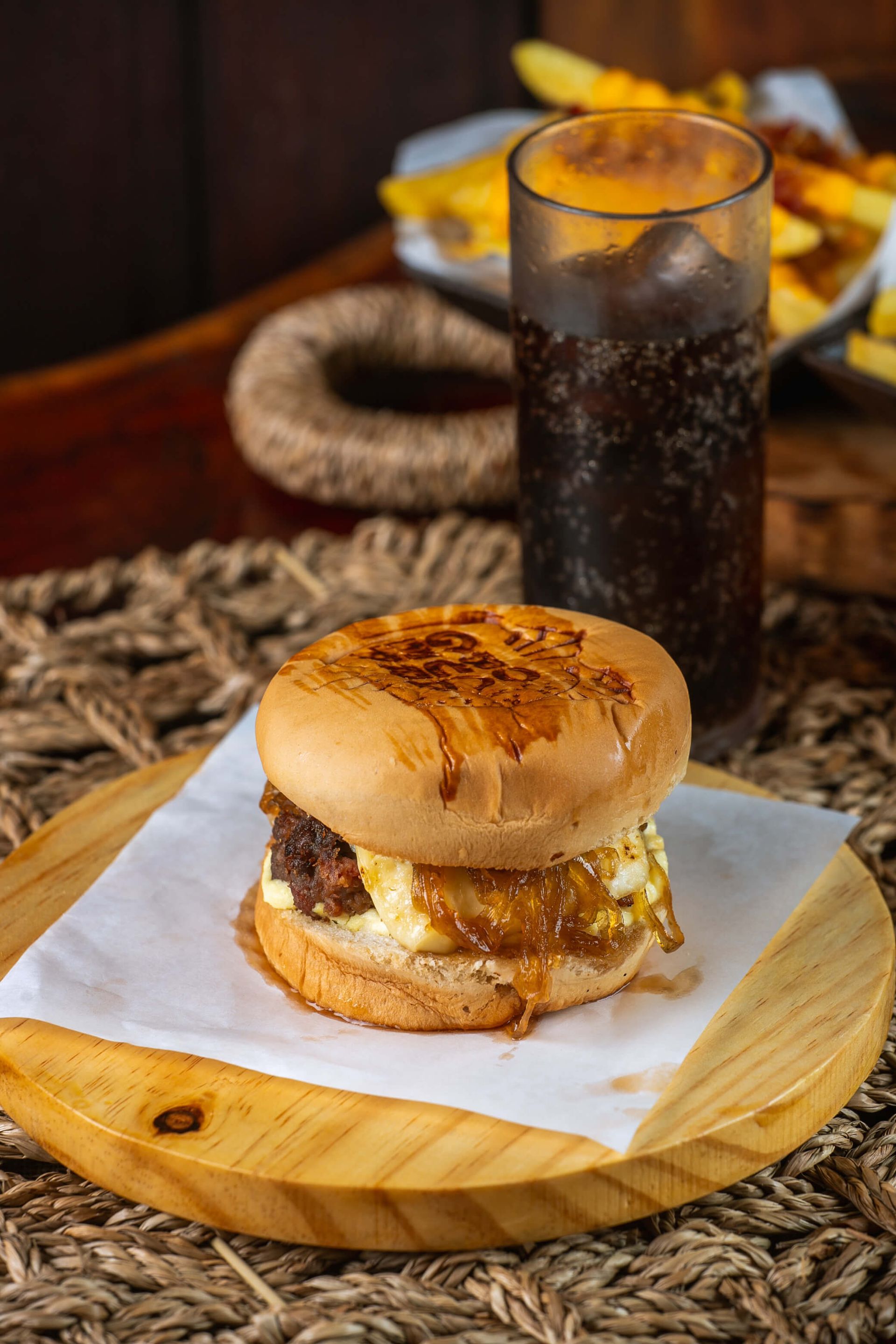 Um hambúrguer está sobre uma tábua de madeira ao lado de um copo de refrigerante.
