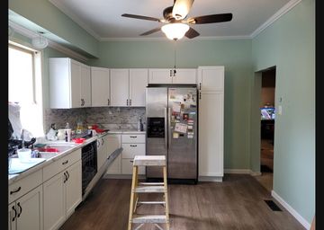 A kitchen with sage green walls, white cabinets, stainless steel appliances, and a central step ladder on wood flooring.