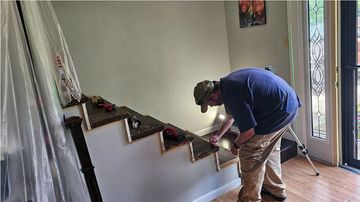 A person in a blue shirt works on the wood trim of an indoor staircase in a brightly lit room.