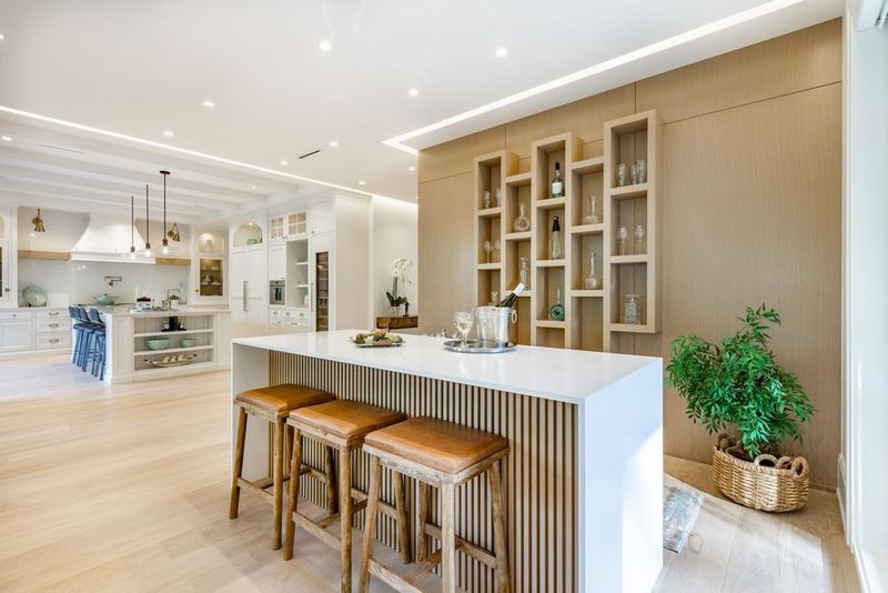 A modern kitchen with a white quartz island, three wooden bar stools, and a light wood-paneled wall with recessed shelving.