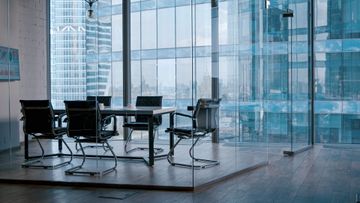 Empty meeting room with four black chairs around a glass table, enclosed by glass walls overlooking city skyscrapers.