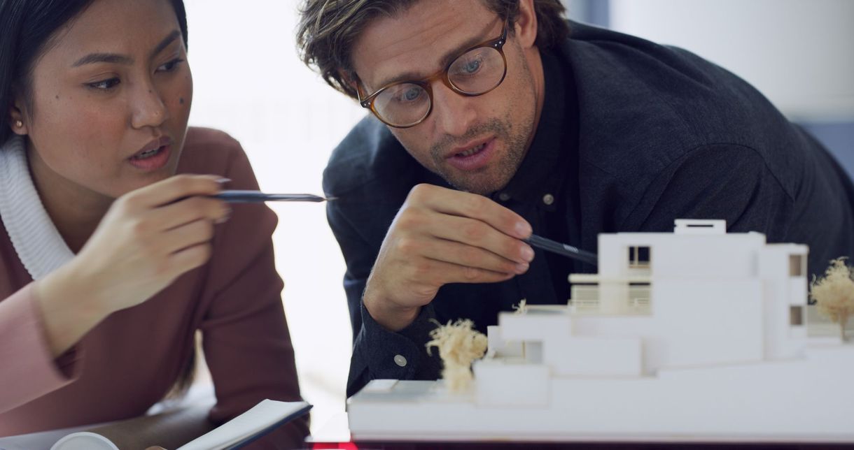 Two professionals examine a white architectural model of a house, pointing with pens while discussing the design.
