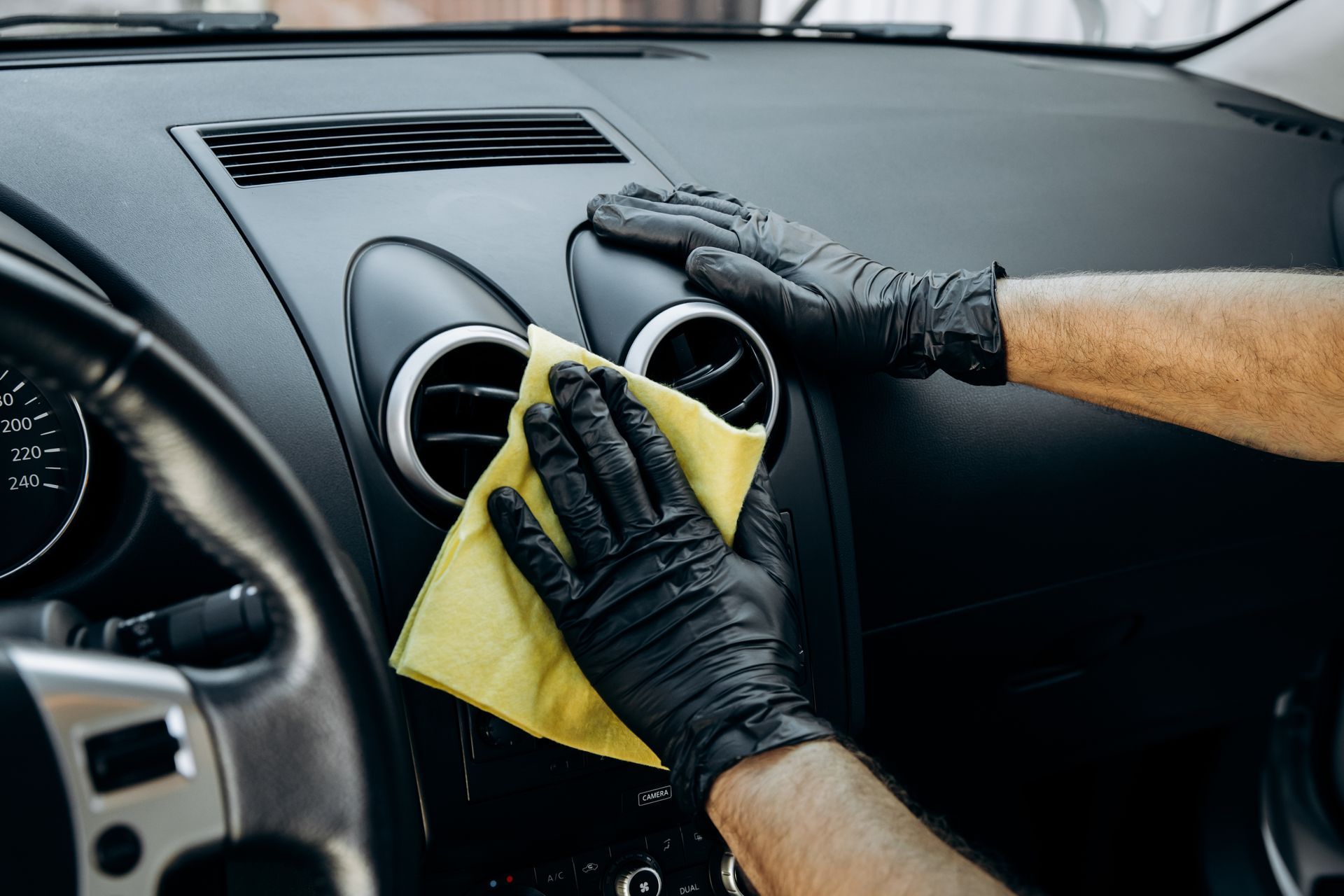 A person wearing black gloves is cleaning the dashboard of a car with a cloth.