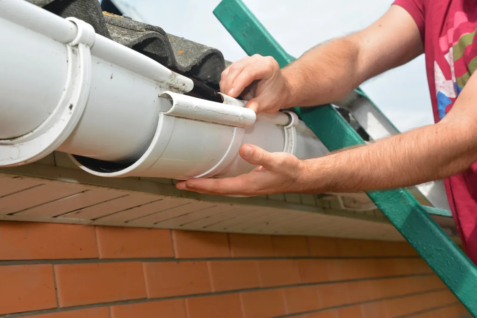 A person installing a white gutter on a brick house, using a green ladder.