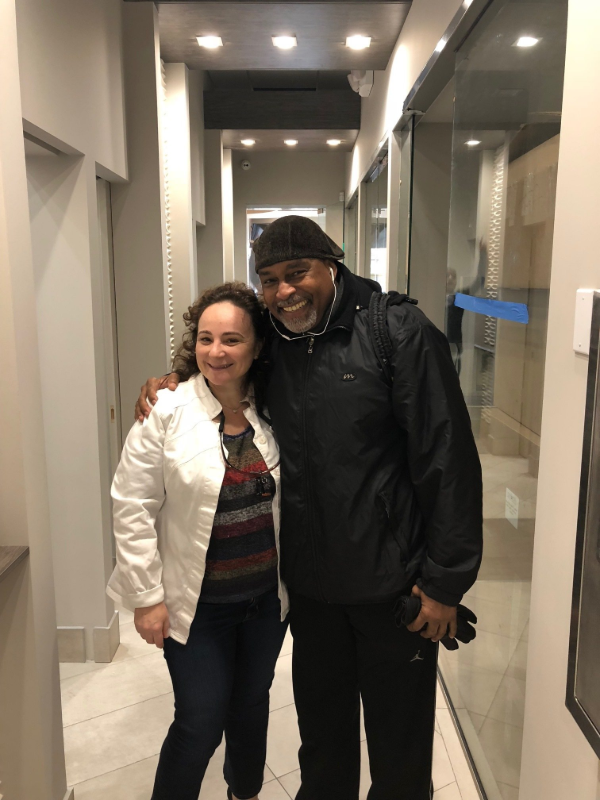 A woman and a man pose smiling in a hallway; she wears a white jacket, he has a black coat and hat.