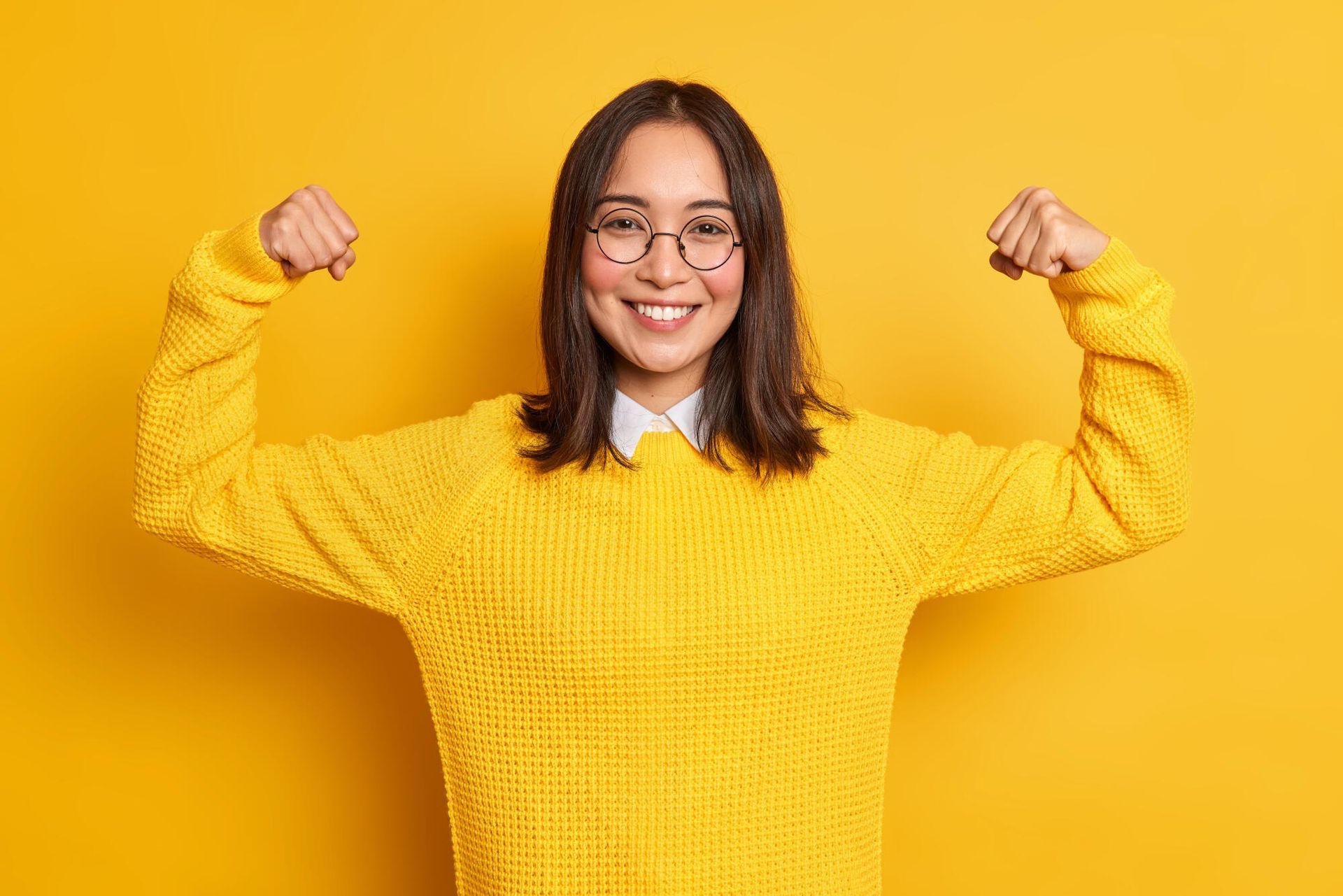 A woman in a yellow sweater is flexing her muscles.