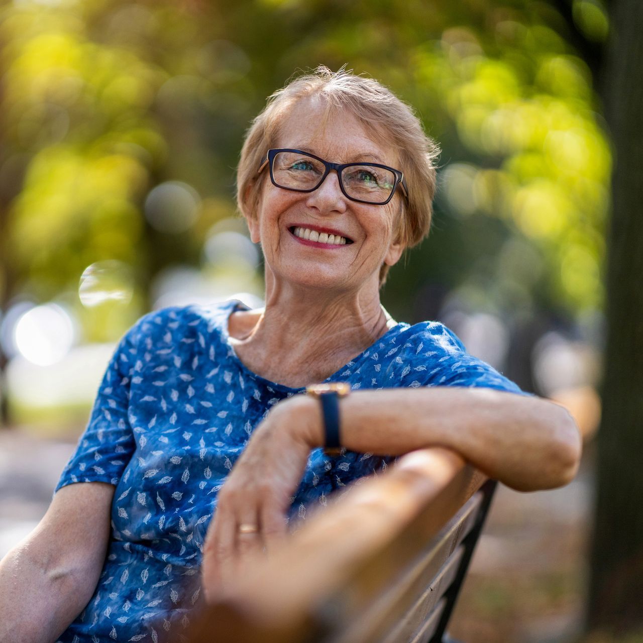 A woman wearing glasses is sitting on a park bench and smiling.