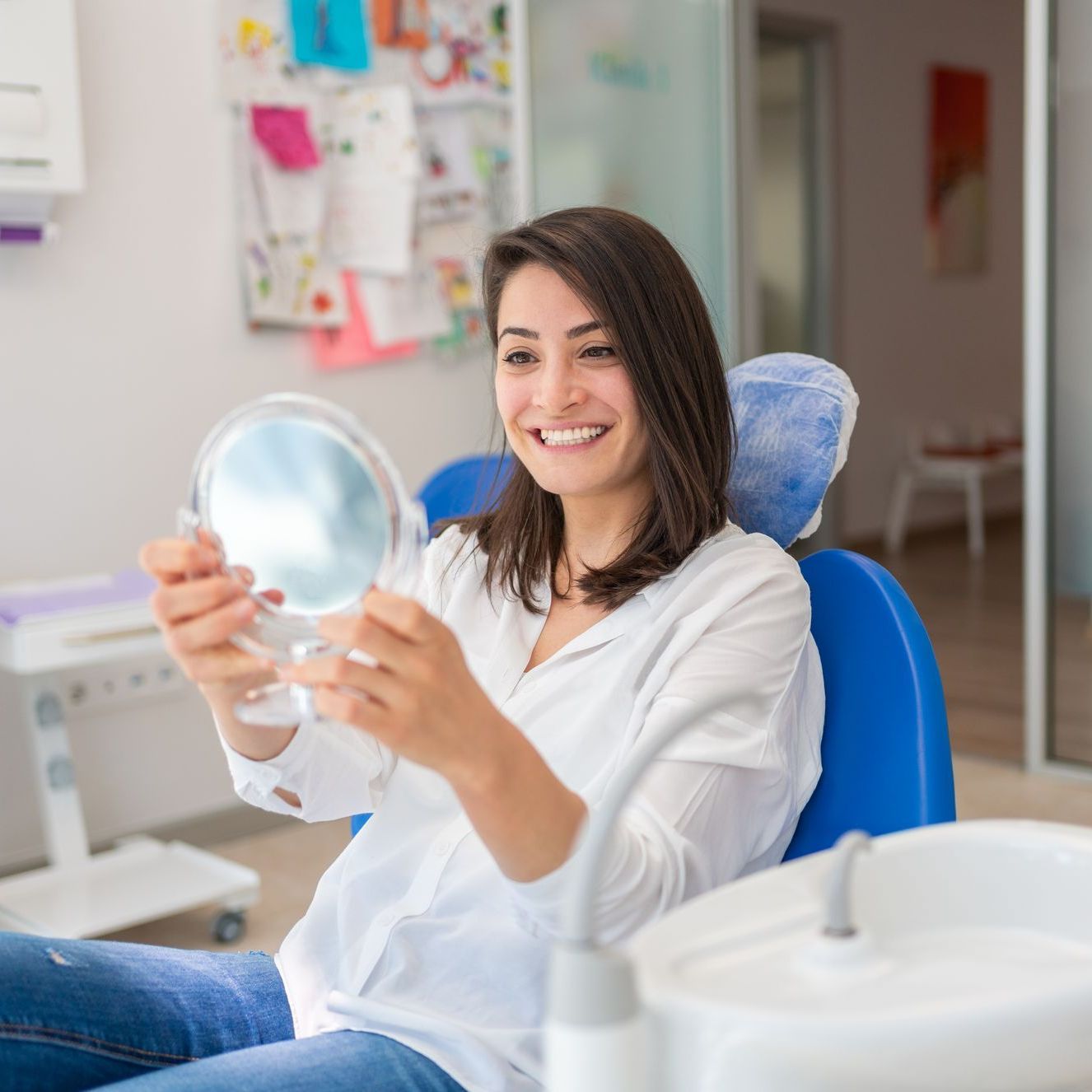 A woman is sitting in a dental chair looking at her teeth in a mirror.