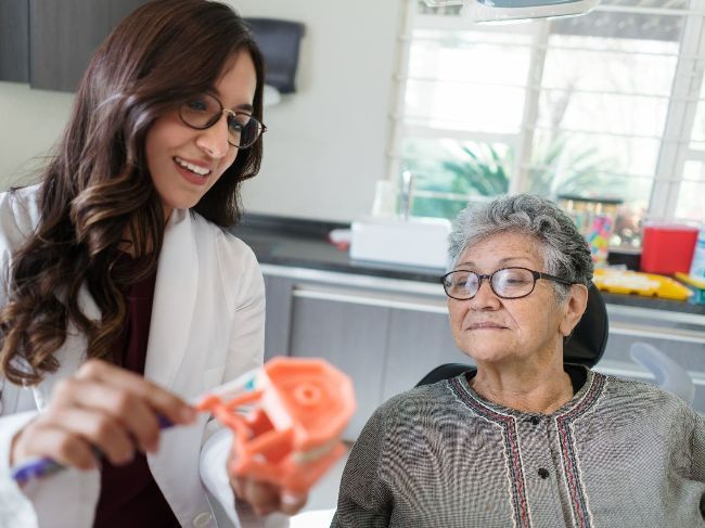 A dentist is showing an elderly woman how to brush her teeth.