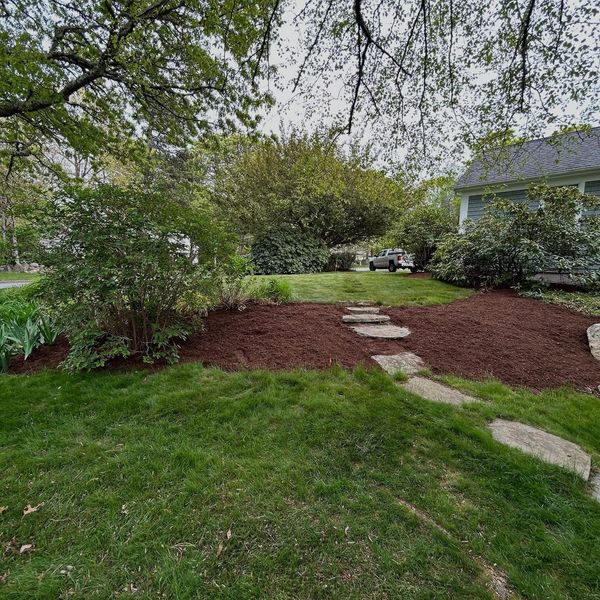 Lawn with mulch beds and stepping stones leading to a building, under a cloudy sky.