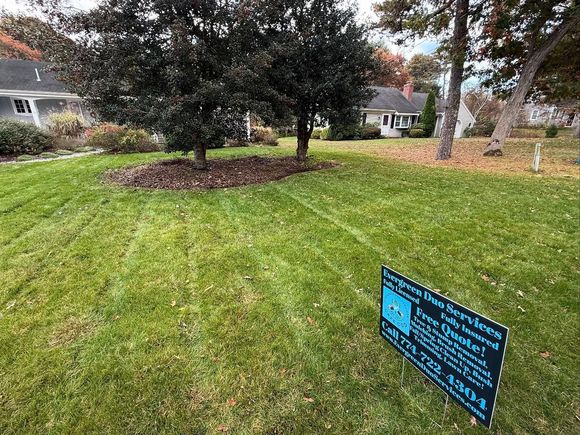 Green lawn with trees, houses in the background, a sign in the foreground with text and phone number.