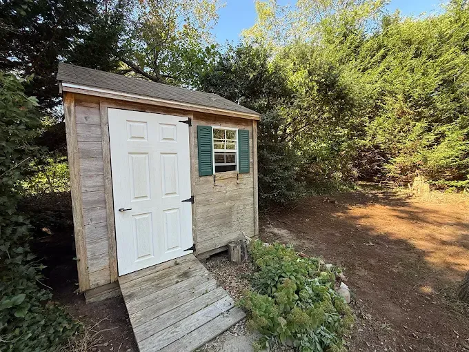 Small wooden shed with white door, green shutters, and ramp, surrounded by trees.