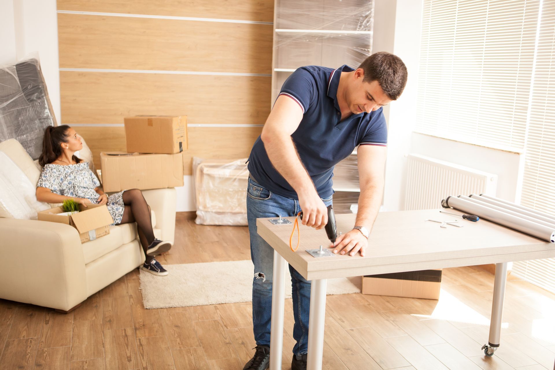 Man assembling furniture, woman resting on a sofa in a room with boxes and blinds.