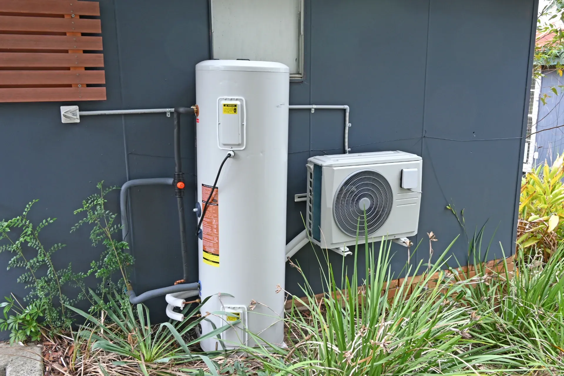 White water heater and outdoor AC unit against a dark exterior wall, with plants in front — New Gen Plumbing and Gas in Banora Point, NSW