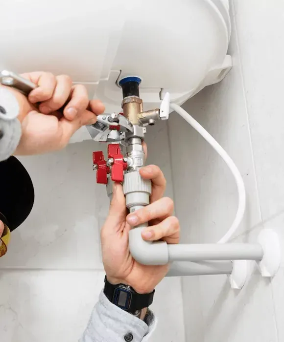 Hands using a wrench to tighten plumbing under a sink with red valve handles and white pipes. — New Gen Plumbing and Gas in Coolangatta, NSW