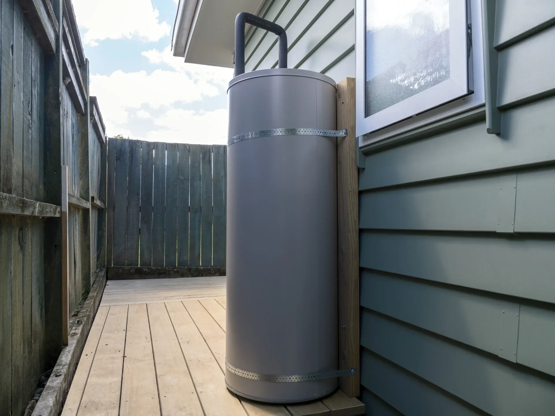 Tall gray cylindrical tank beside a house wall on a narrow wooden deck between fences — New Gen Plumbing and Gas in Banora Point, NSW