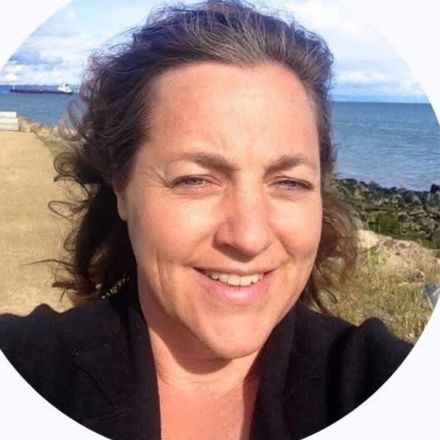 Woman with curly brown hair smiling outdoors, ocean and sky in background.