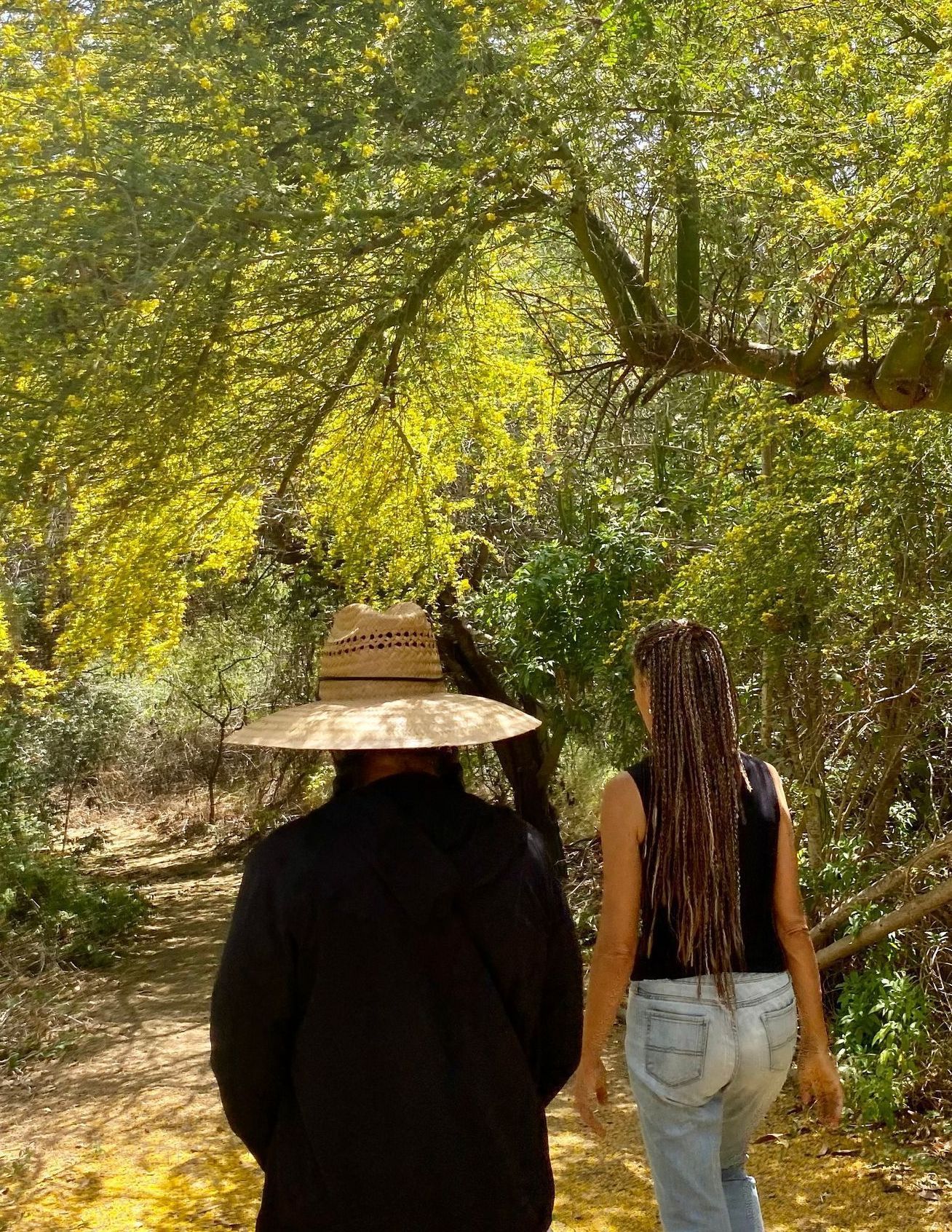 Two people walking on a path in a wooded area, one wearing a hat and black jacket.