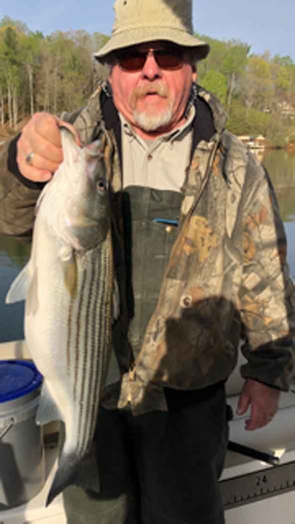 Man holding a large striped bass on a boat, wearing camo jacket and hat, with water and trees behind him
