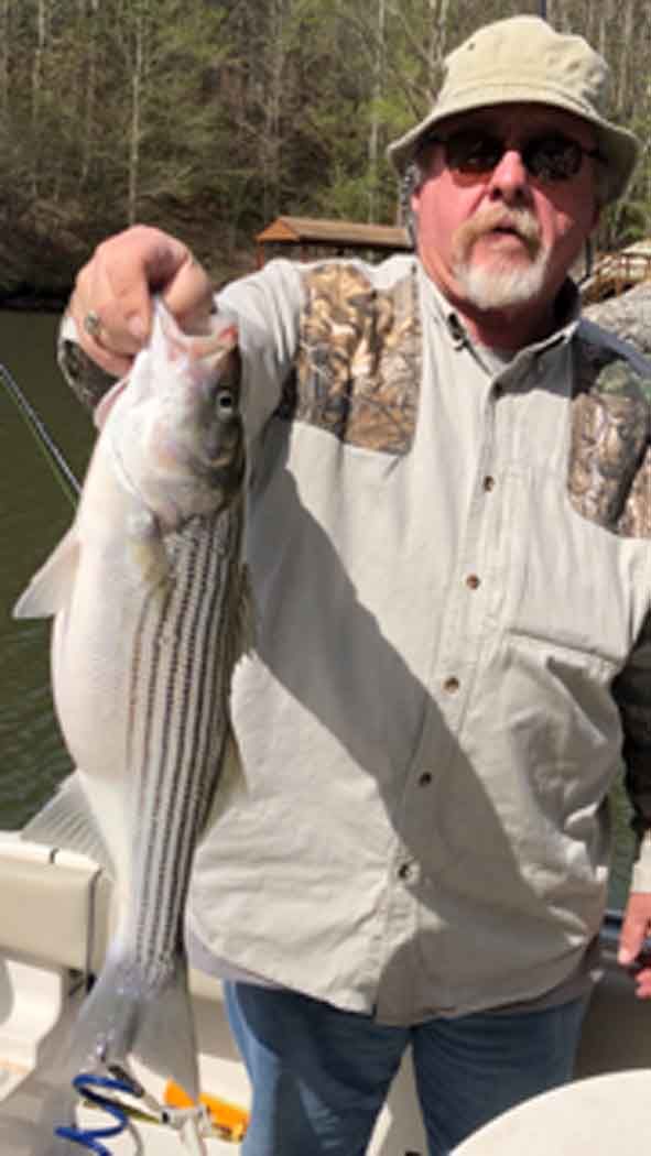 Man holding a striped fish on a boat, wearing a light shirt, hat, and sunglasses.
