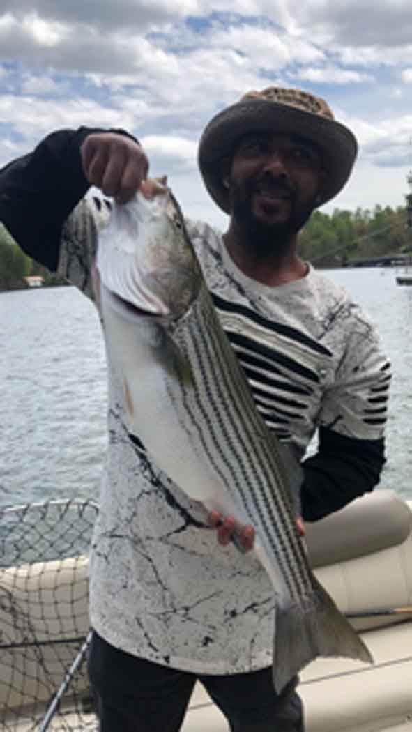 Person holding a large striped fish by the water on a boat, smiling at the camera.