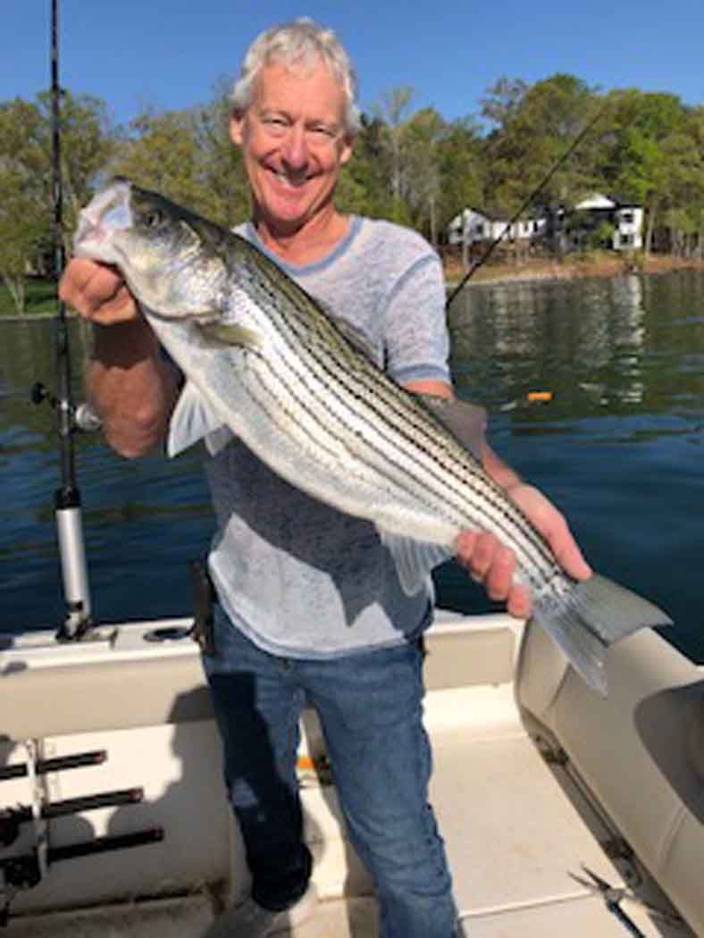 Man holding a large striped fish on a boat, smiling on a calm lake with trees in the background