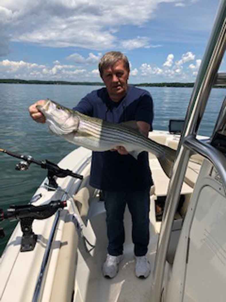 Man standing on a boat holding a large striped fish over the water under a partly cloudy sky