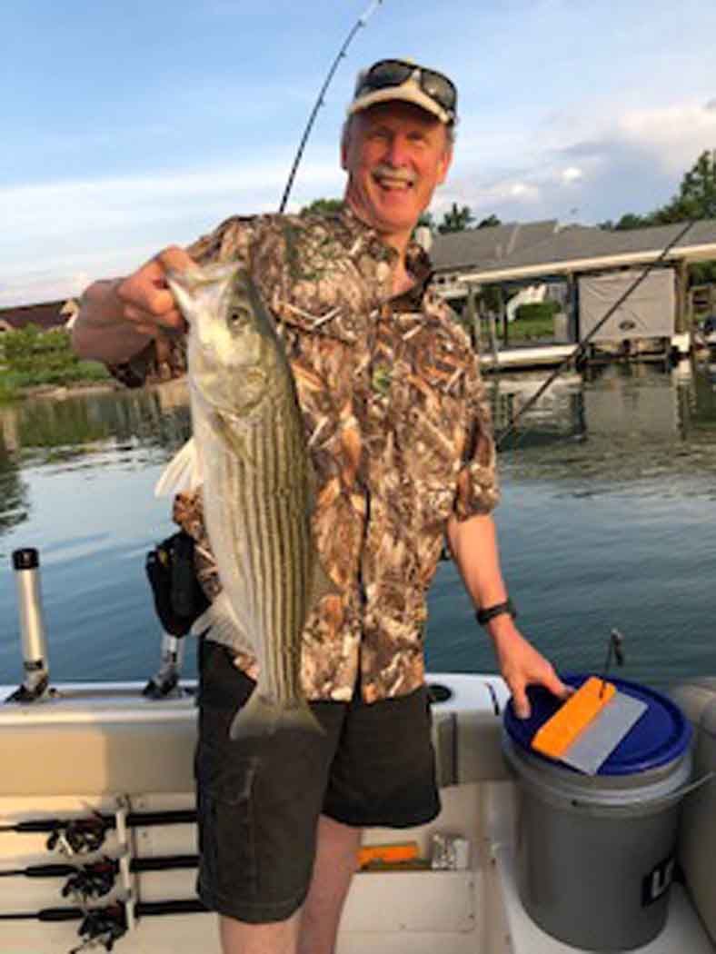 Angler holding a large striped bass on a boat at sunset near a dock