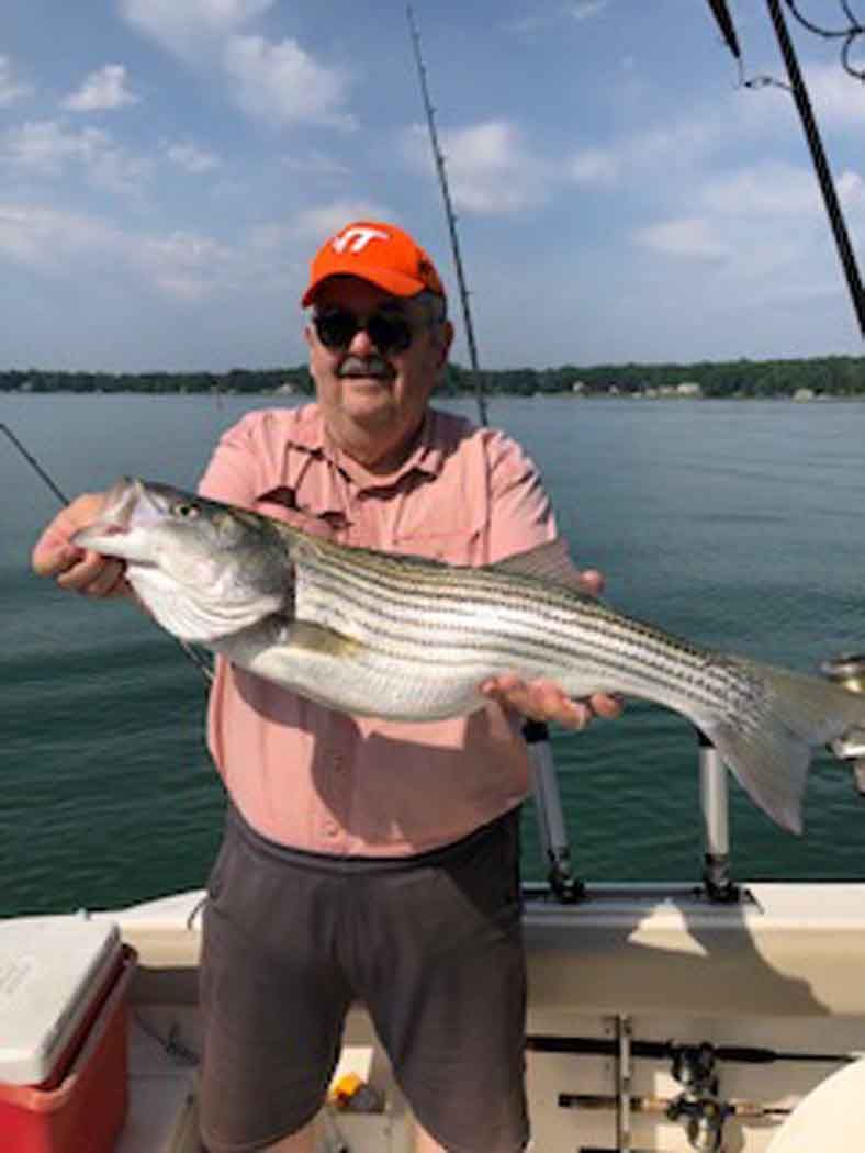 Man holding a striped fish on a boat, with calm water and shoreline behind him.