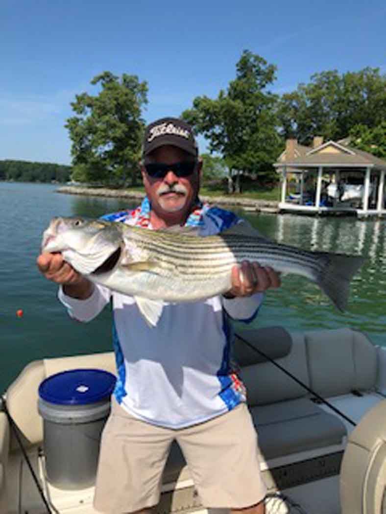 Man holding a large striped fish on a boat beside a calm lake and dock under a blue sky