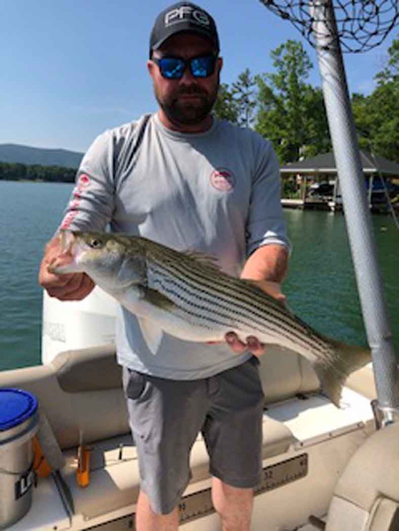 Man holding a large striped fish on a boat by a lake on a sunny day