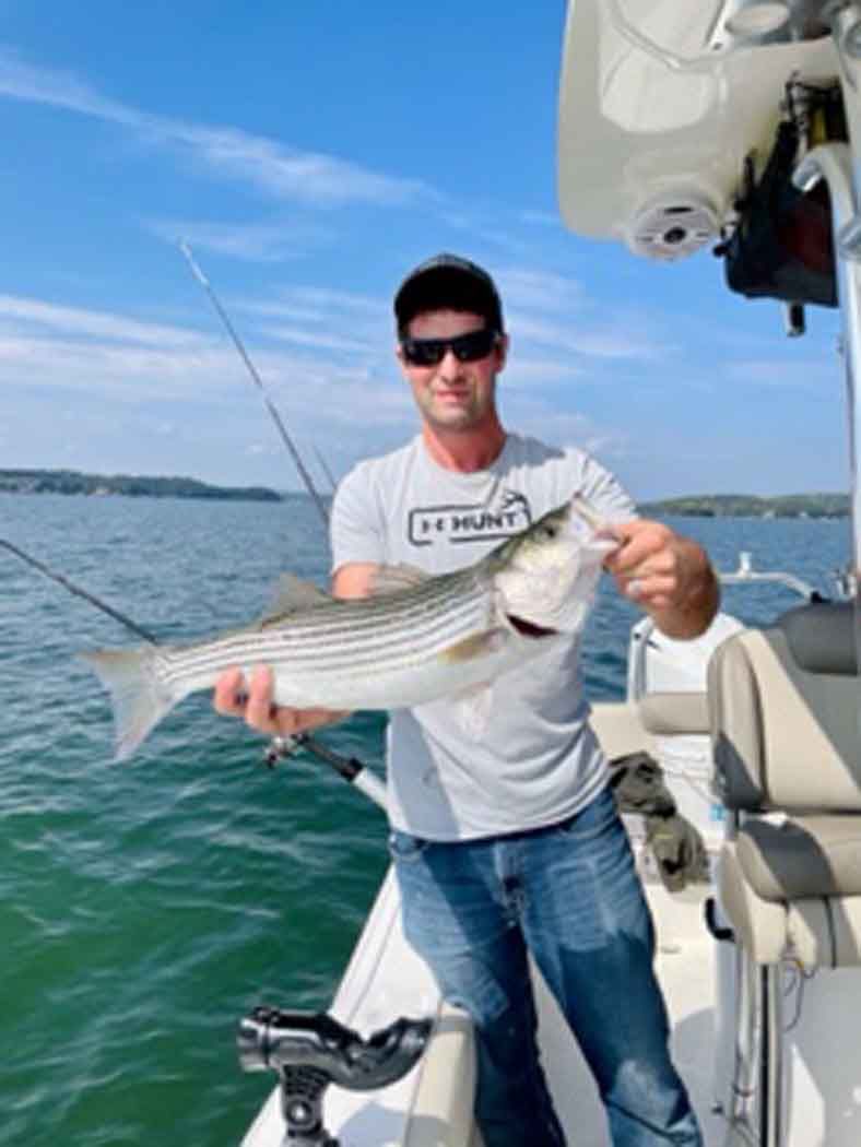 Man holding a striped fish on a boat on calm blue water under a clear sky