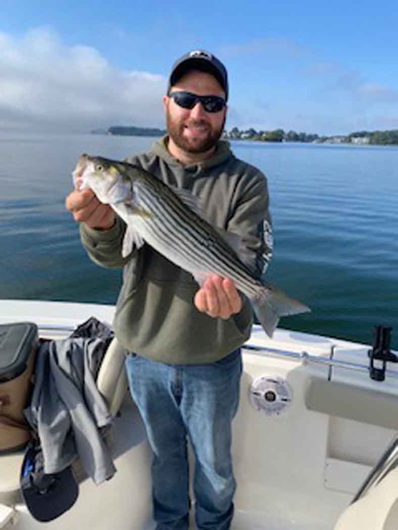 Man holding a large striped fish on a boat, with calm water and distant shoreline behind him.