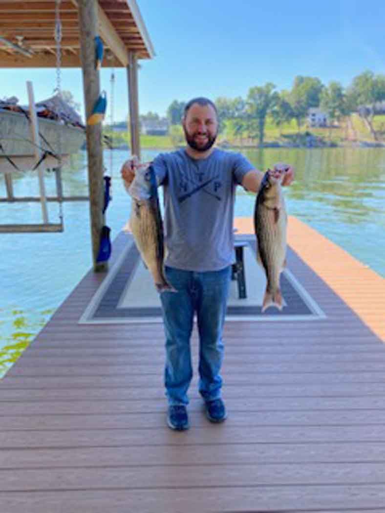 Man standing on a dock holding two large fish, with water and trees in the background.