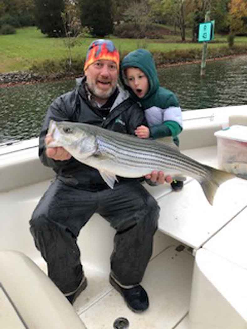 Man and child on a boat holding a large striped fish by a river