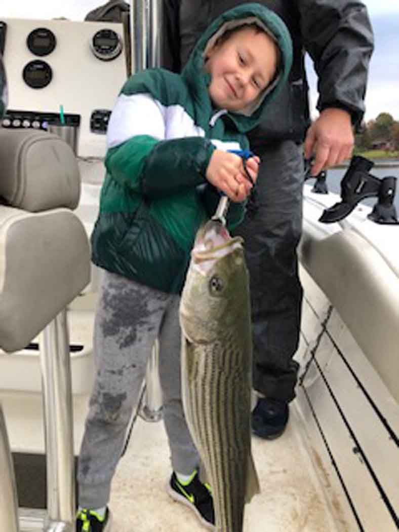 Child smiling while holding a large striped fish on a boat, with an adult standing behind.