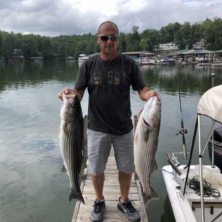 Man holding two large striped fish on a dock beside a lake, with a boat and trees in the background