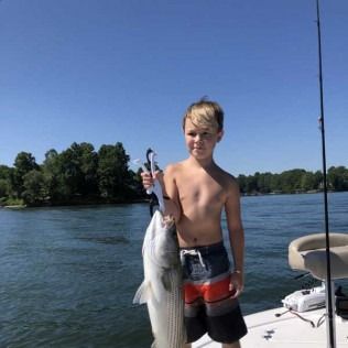 Boy on a boat holding a large fish on a lake, with trees and blue sky in the background.