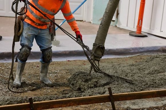 Construction worker pours concrete with a hose, wearing orange safety gear, knee pads, and rubber boots.