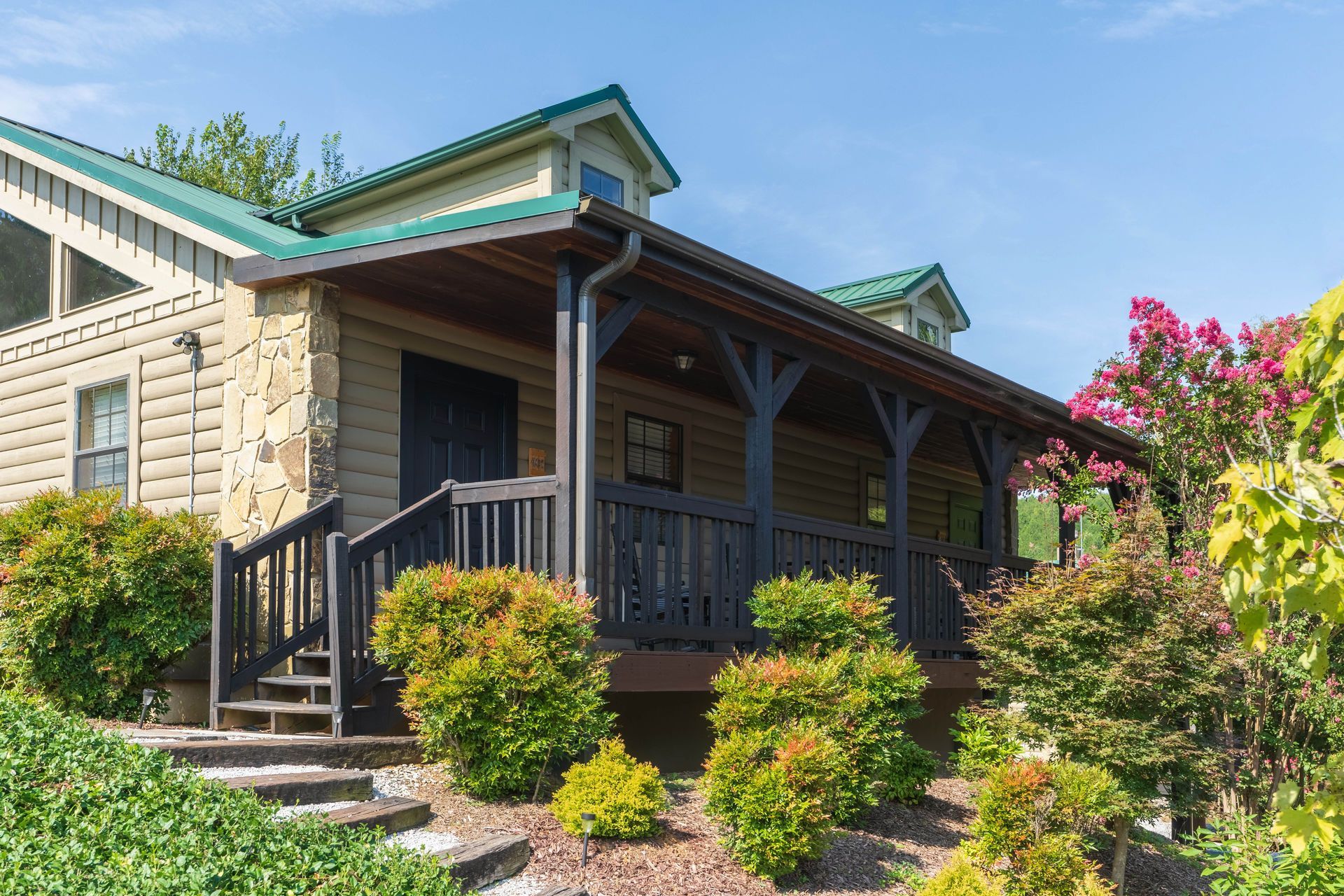 Cabin with a green roof and a porch, surrounded by green bushes and flowering pink tree.