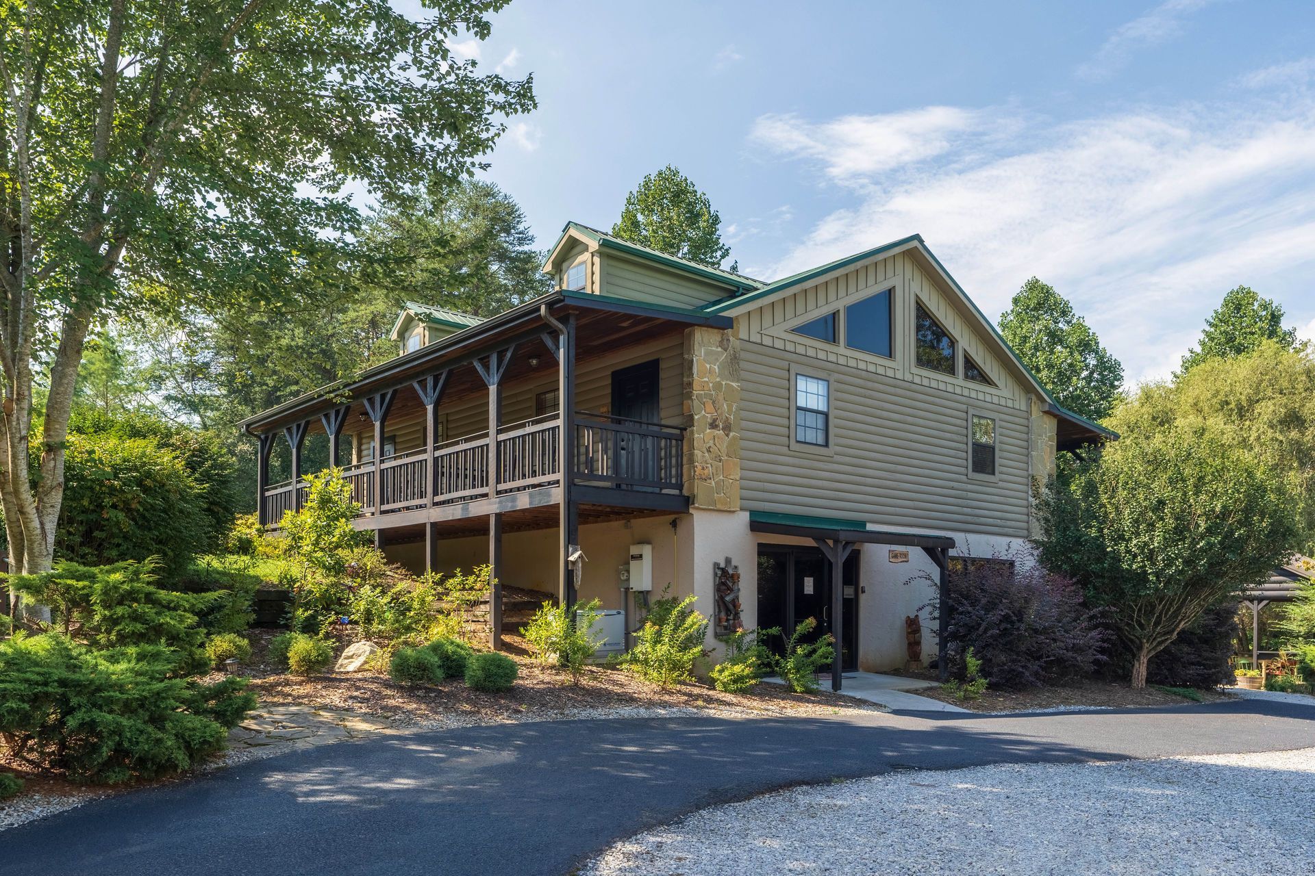 Two-story cabin with a long porch, stone and wood siding, trees in the background, and a gravel driveway.