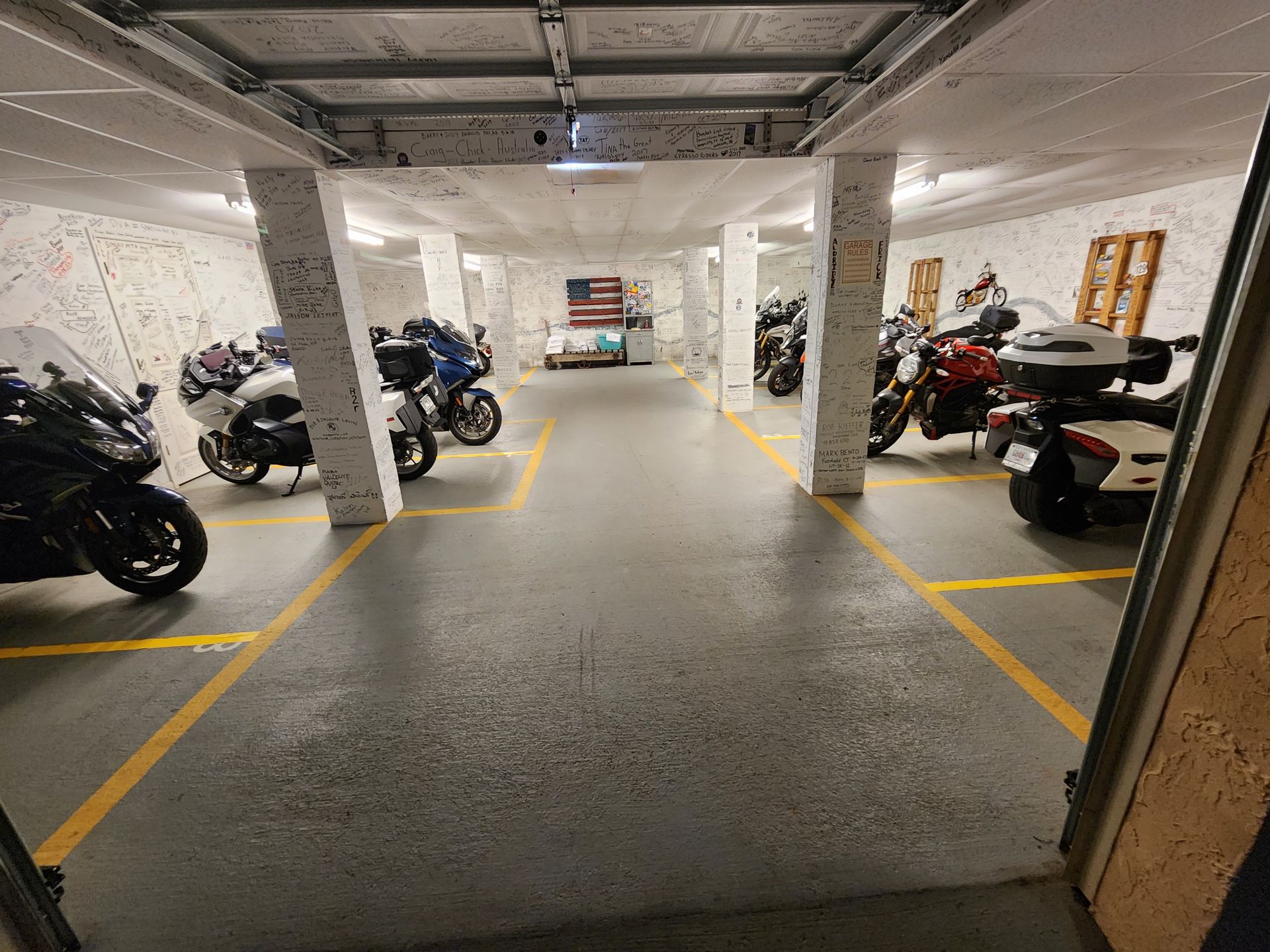 Motorcycles parked in a garage with yellow parking lines, under a concrete ceiling, and a US flag on the back wall.