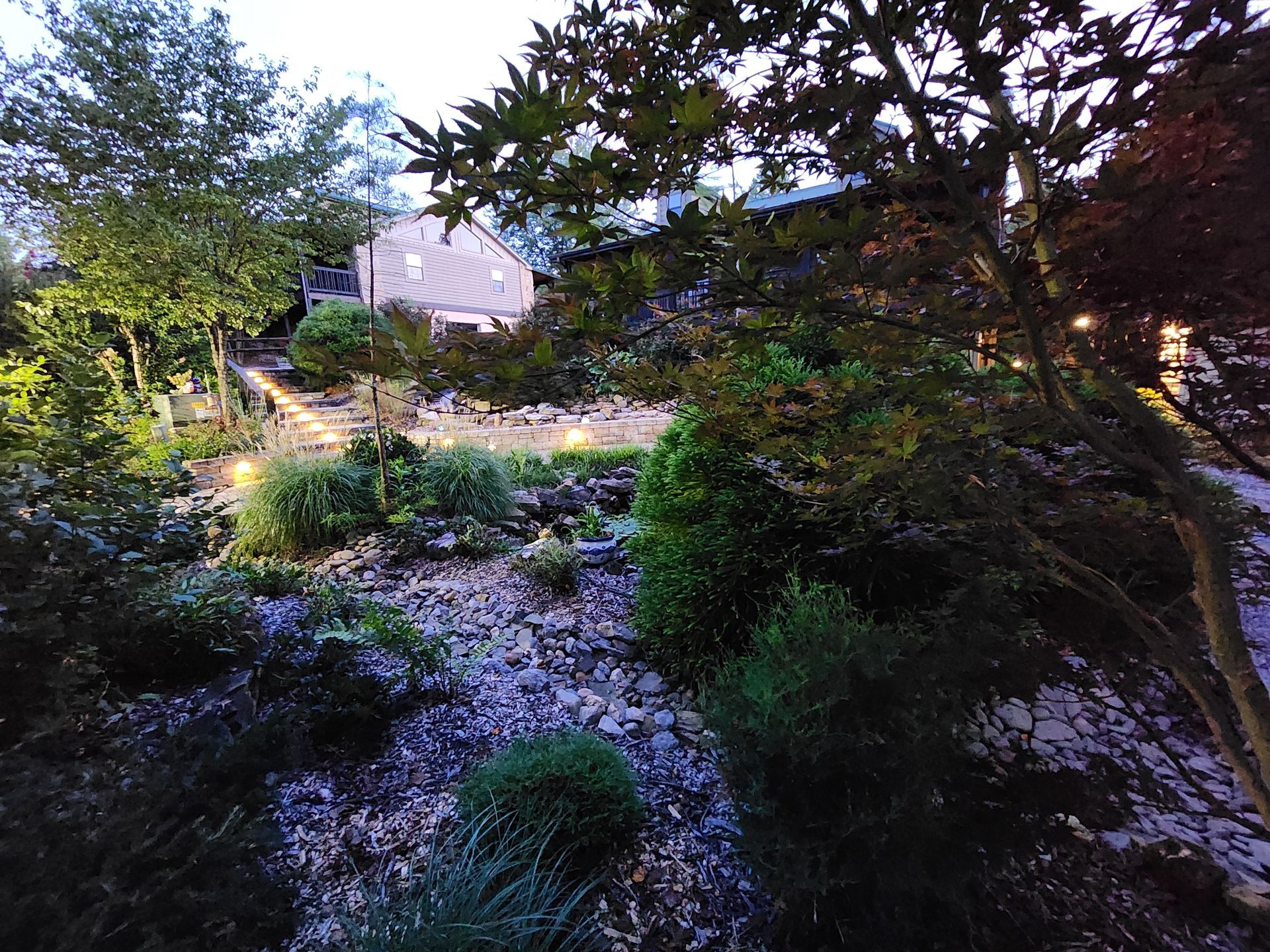 Landscaped garden at dusk, illuminated by lights; a building is visible in the background, surrounded by trees and shrubbery.